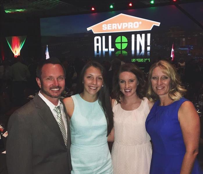 Man and three women standing in a dark room with a SERVPRO orange house logo behind them with the words "All In!" 